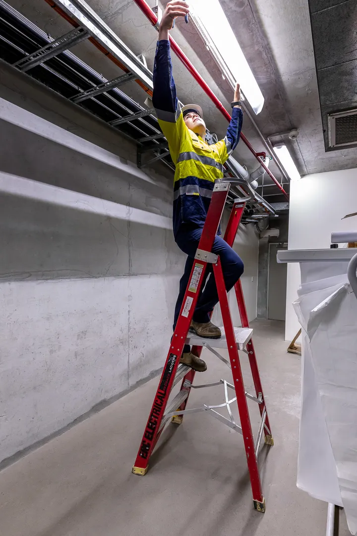 Worker on ladder repairing overhead light fixture in warehouse.