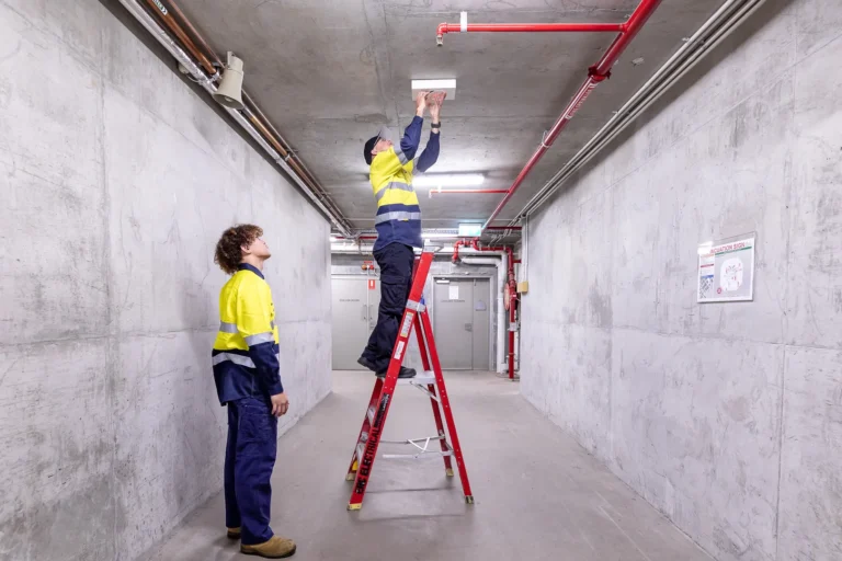 Workers on ladder inspecting ceiling pipes.